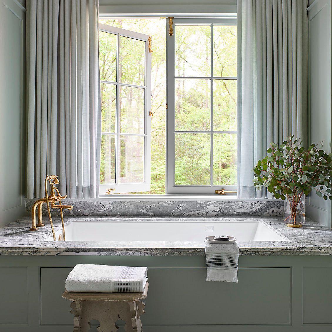 Bright kitchen sink beneath a window with open shutters, framed by pale curtains and a potted plant.