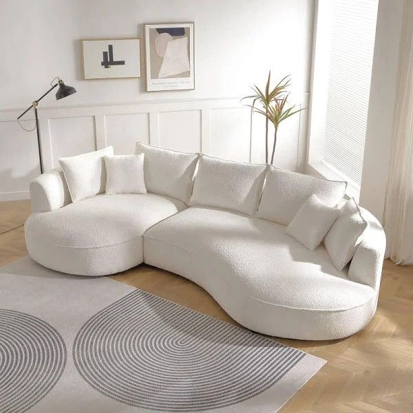 Modern white curved sofa in a bright minimalist living room with wall art, lamp, plant, and patterned rug