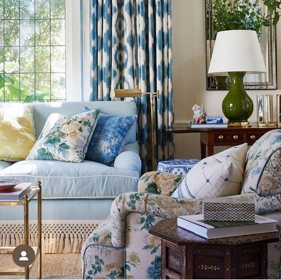 Bright living room with blue floral sofas, patterned pillows, a lamp, and large windows with greenery outside