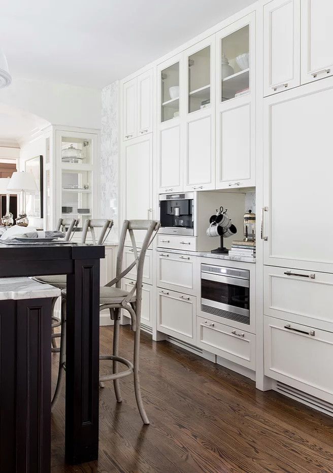Bright white kitchen with built-in cabinets, stainless oven, and dark wood floor, viewed from the island