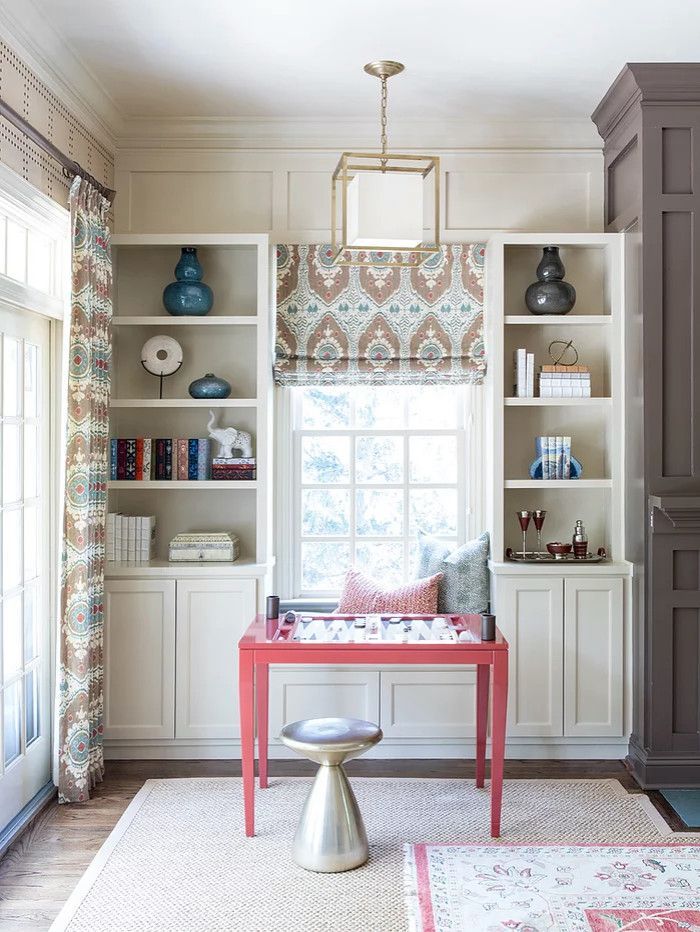Built-in home office with cream shelves, coral desk, patterned window shade, and stool by a bright window