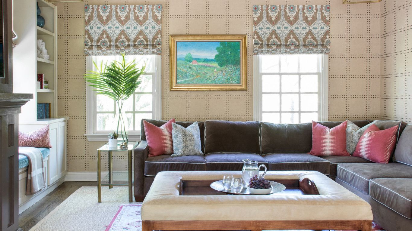 Bright living room with a gray sectional, pink pillows, coffee table, and framed art by patterned windows