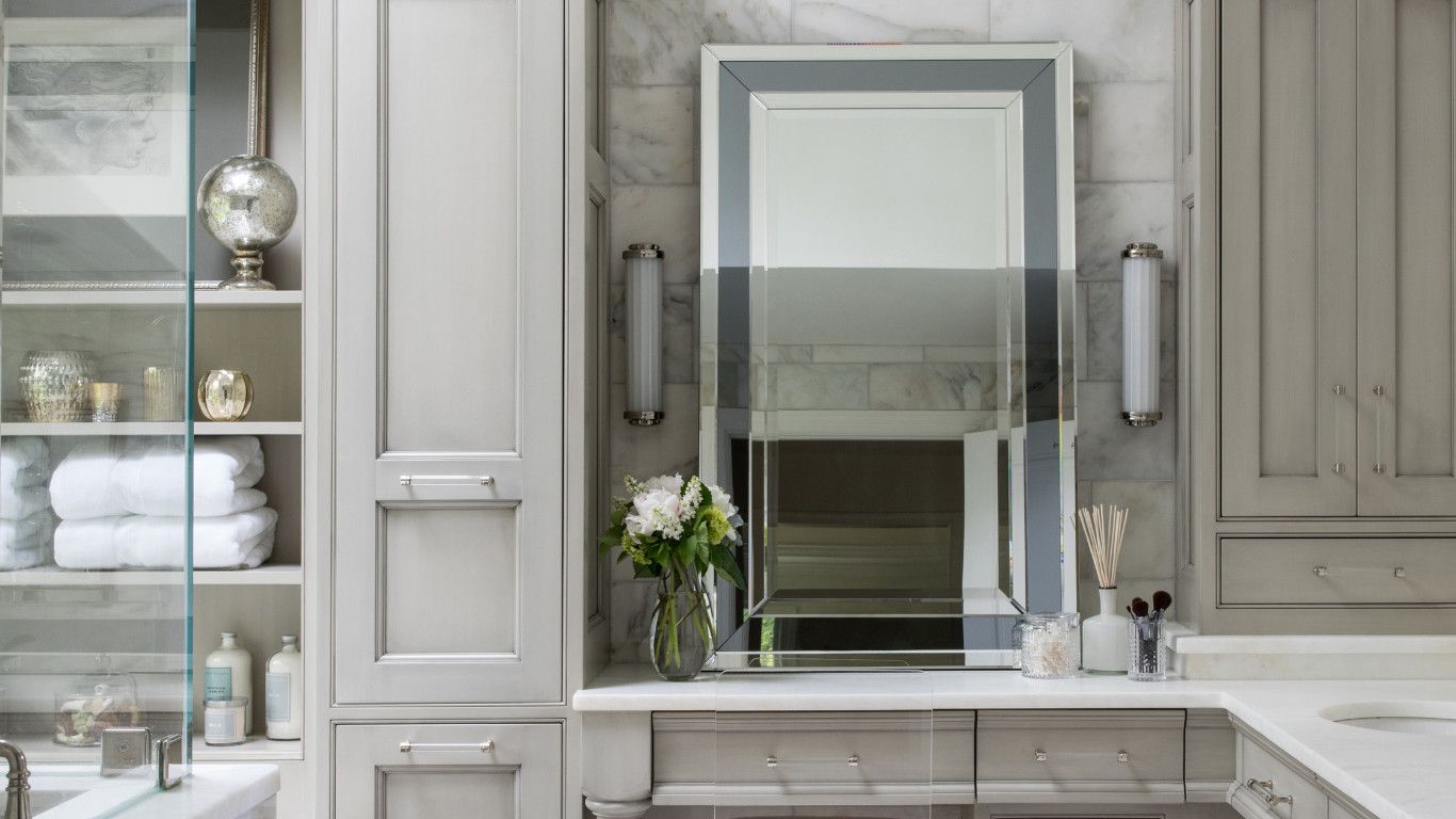 Elegant bathroom vanity with a centered mirror, gray cabinets, shelves of towels, and white flowers on the counter.