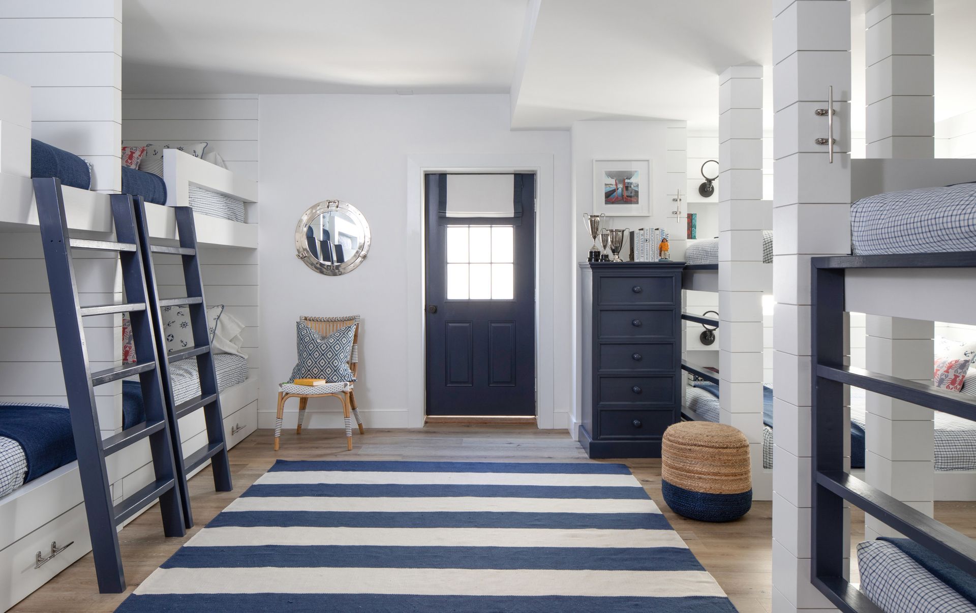Bright blue-and-white bedroom with bunk beds, striped rug, navy door, and a small dresser and chair.