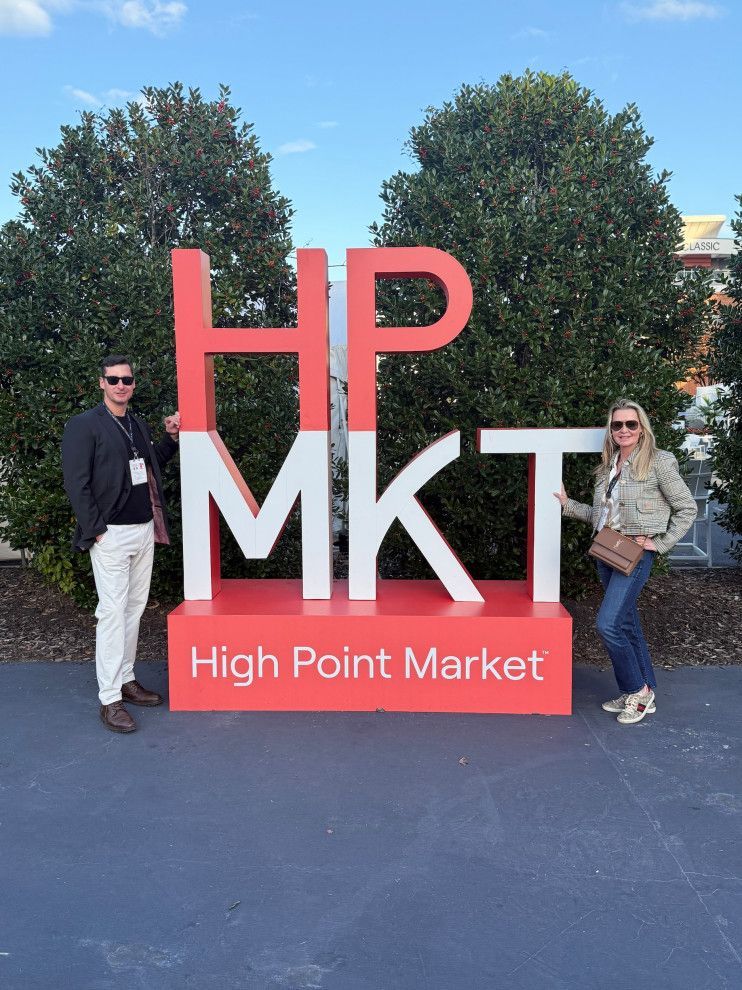 Two people posing beside a large red and white “HP MKT” High Point Market sign outdoors
