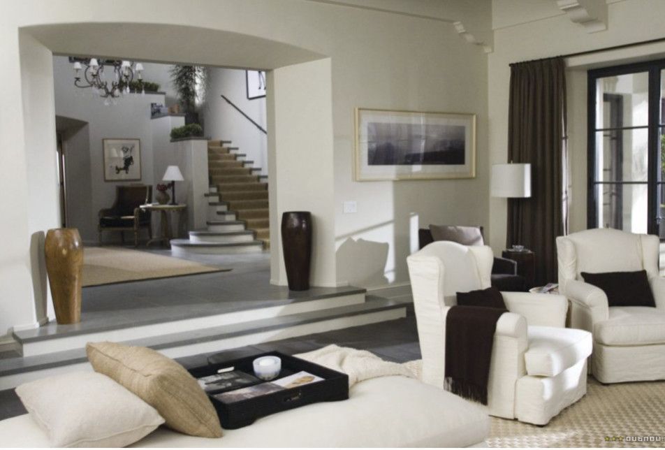 Modern living room with white sofas, dark accent chairs, staircase, and large mirror opening to another room