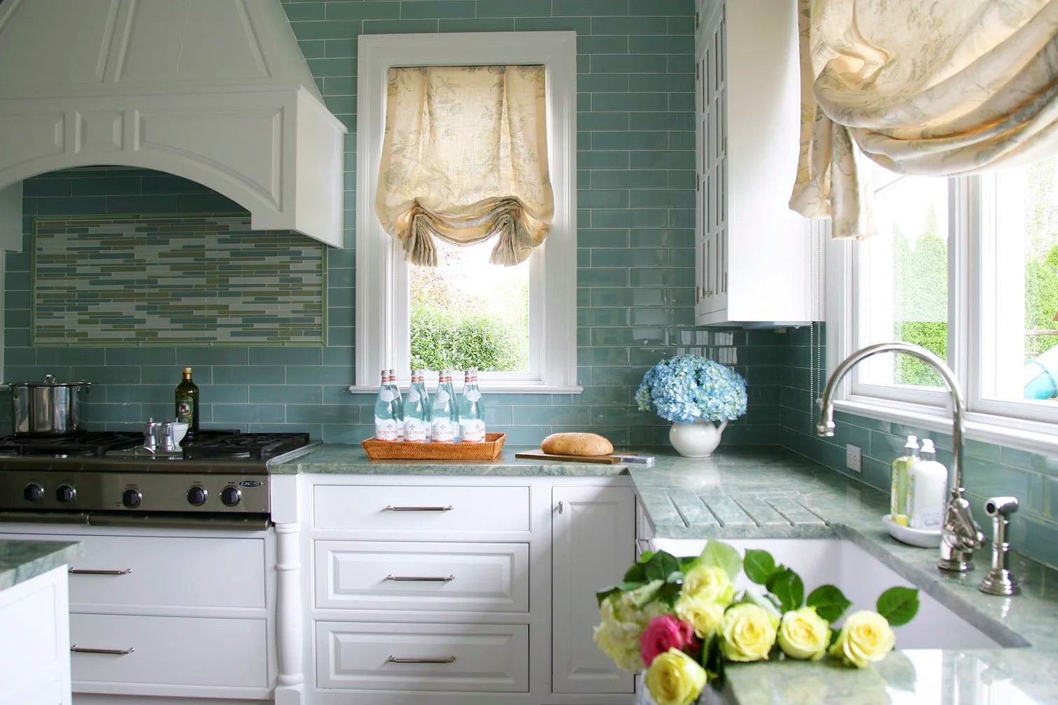 Bright kitchen with teal tile, white cabinets, sunlit windows, and flowers on the counter