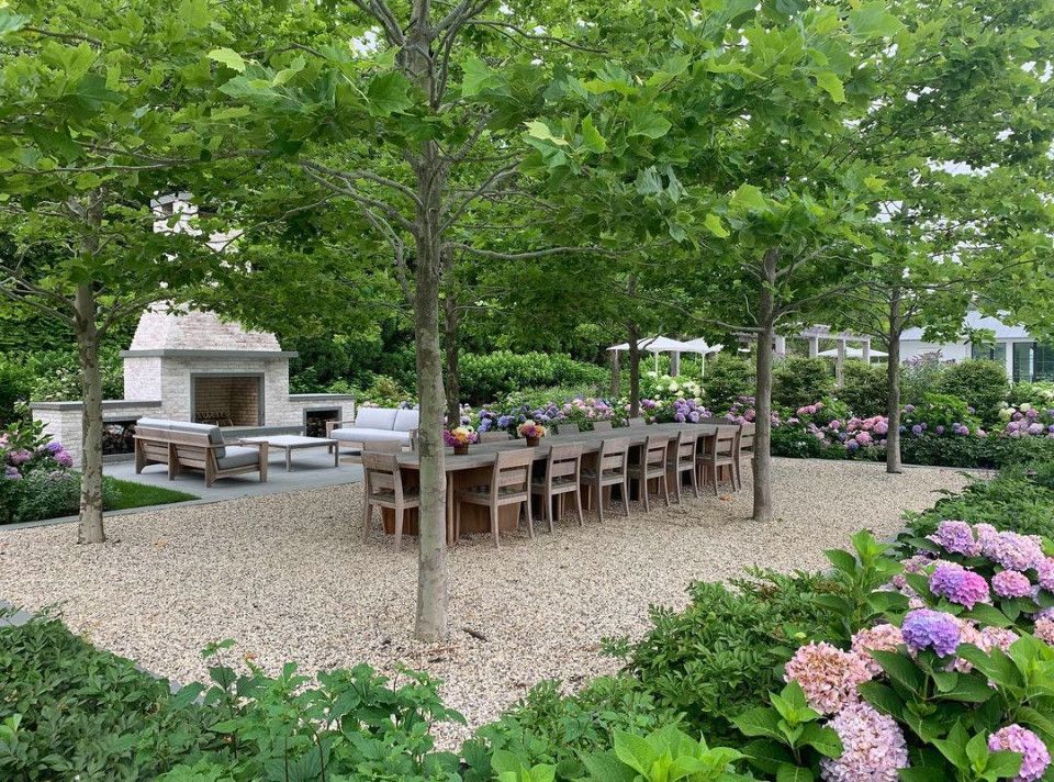 Outdoor patio dining area with long wooden table, grill, and pink hydrangeas under green trees