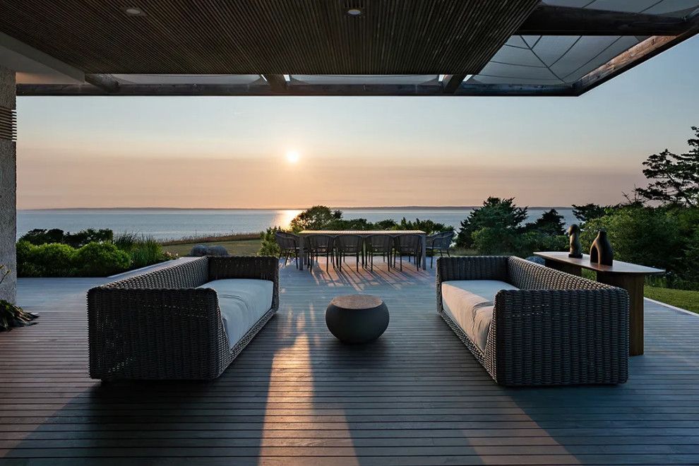 Modern patio lounge overlooking a calm ocean at sunset, with wicker seating and a fire pit