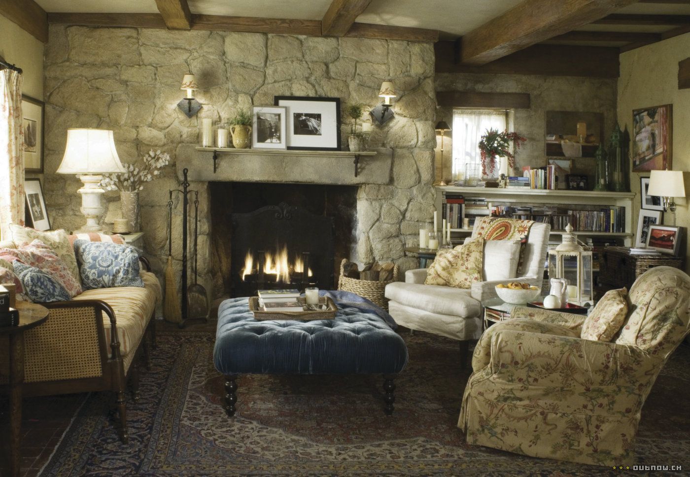 Cozy stone living room with a lit fireplace, armchairs, lamp, and blue tufted ottoman.