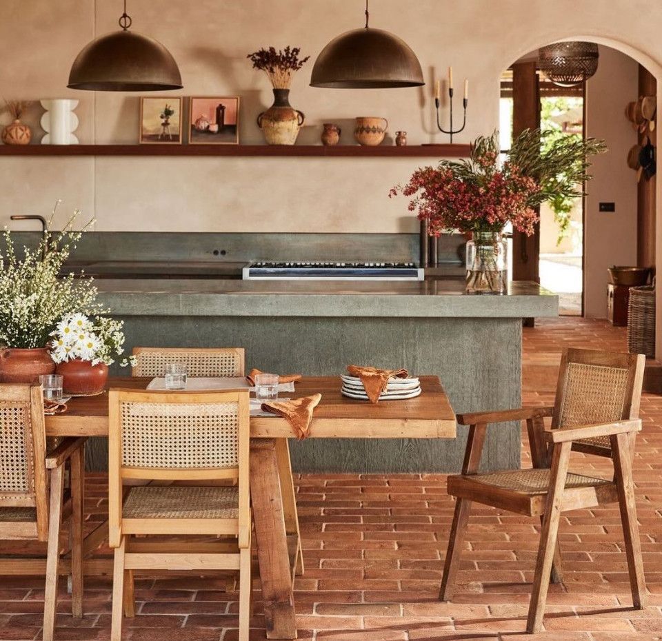 Warm rustic kitchen with stone island, woven chairs, terracotta floor, and a sunlit arched doorway
