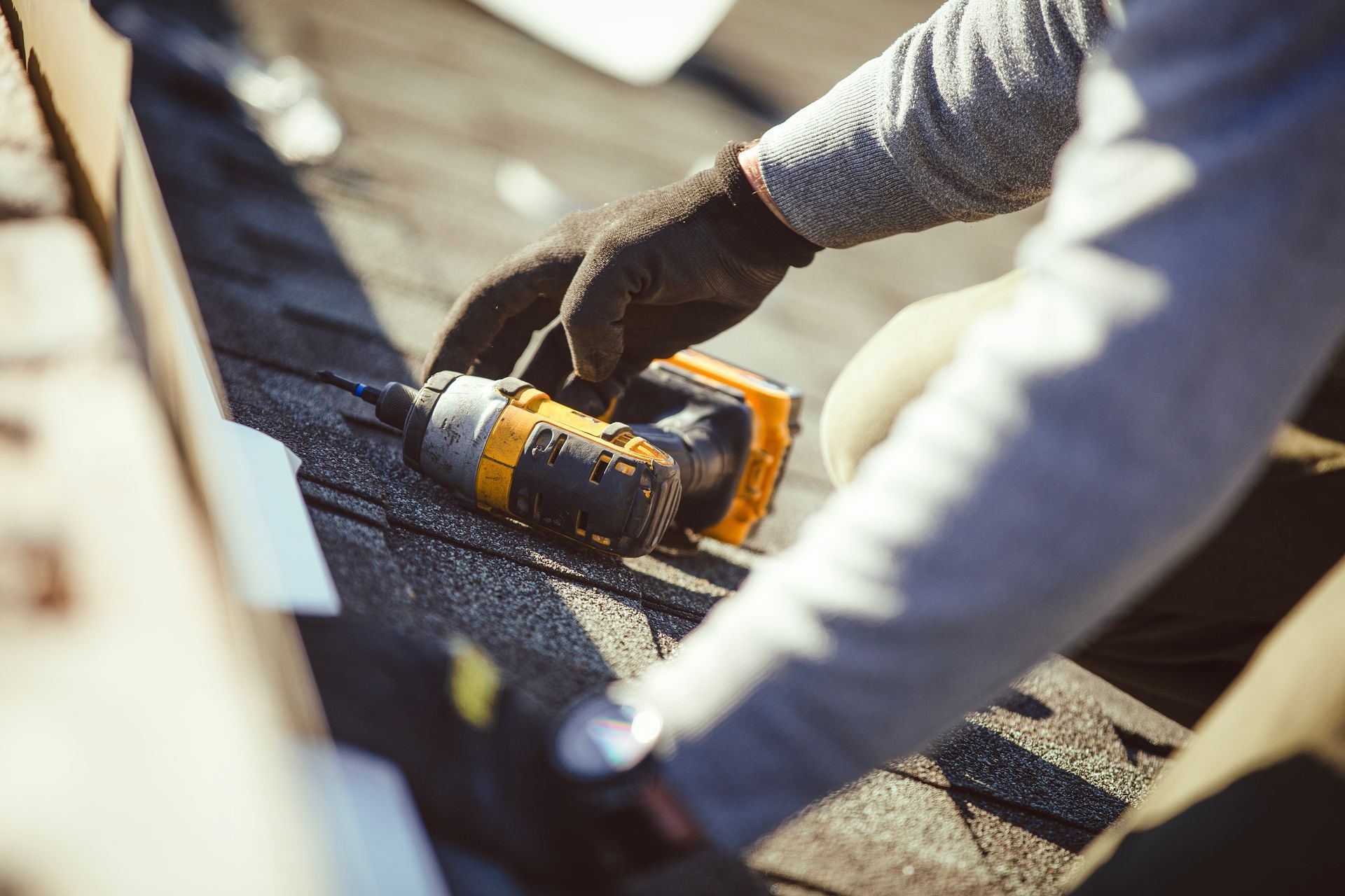Close-up of gloved hands using a power drill to repair roof shingles.
