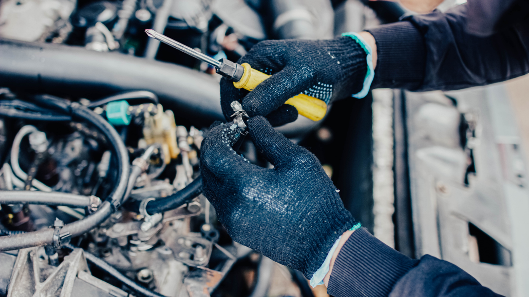 Gloved hands working on car engine wiring with a yellow tool in an auto repair shop