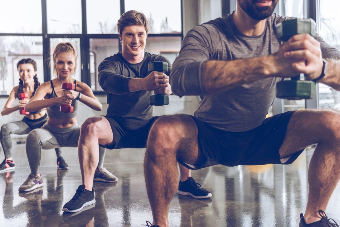 Lezione di fitness di gruppo in palestra con esercizi di squat, con partecipanti che tengono in mano dei manubri e un istruttore davanti.