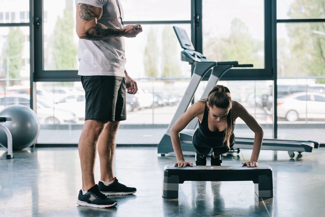 Un uomo e una donna si allenano in palestra; la donna fa flessioni su una panca mentre l'uomo è in piedi accanto a lei.