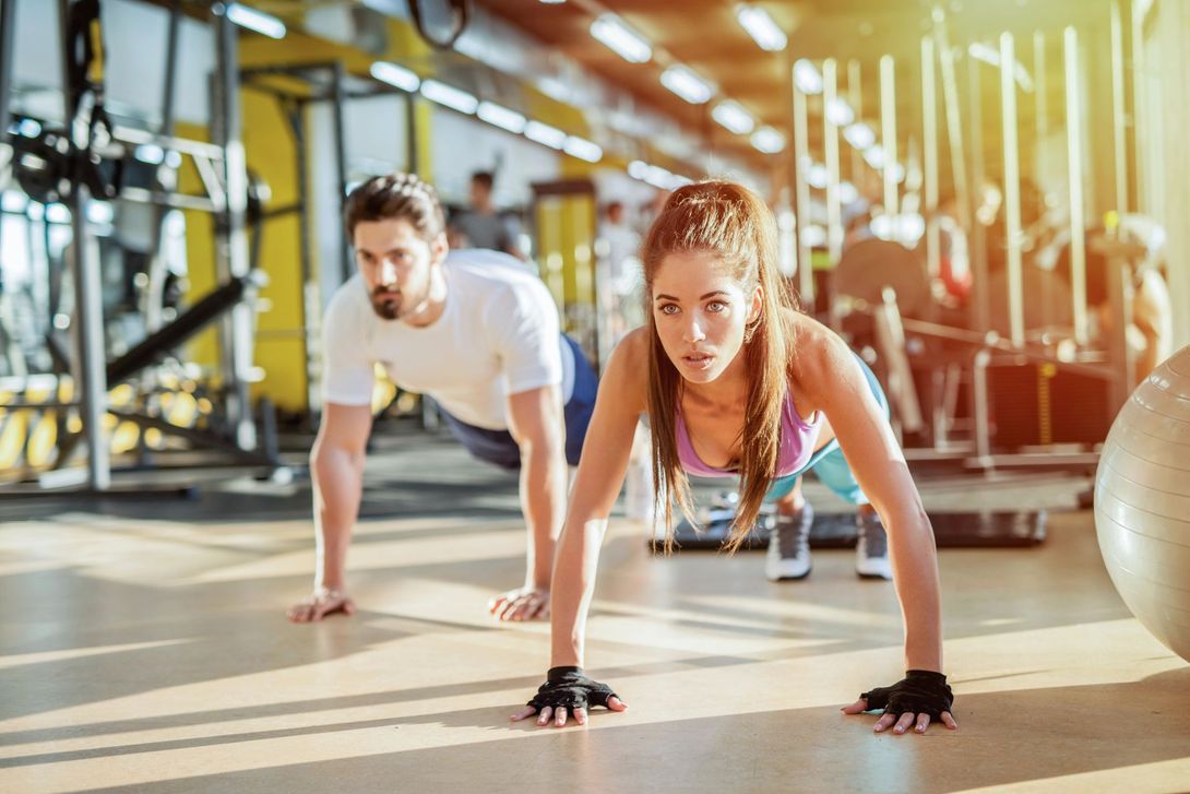 Due persone che fanno flessioni in una palestra luminosa, con attrezzi ginnici sullo sfondo.