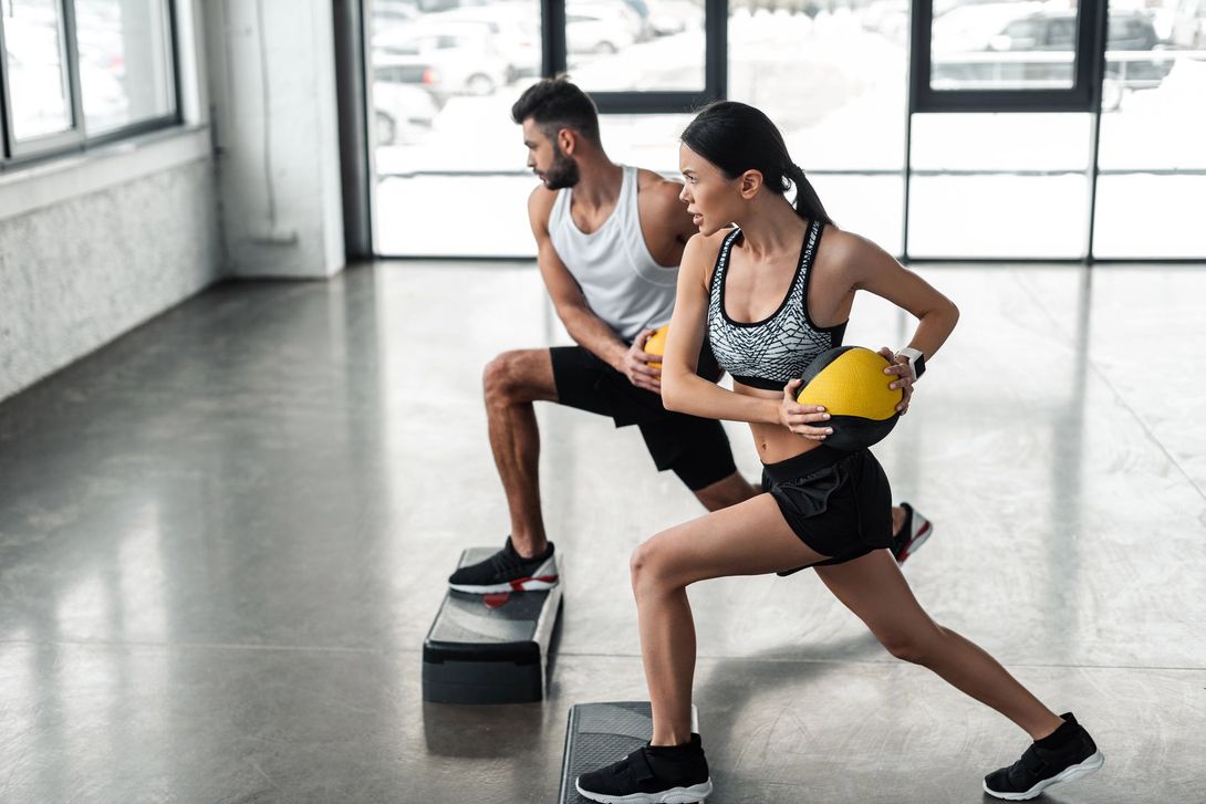 Due persone che fanno step-up in una palestra luminosa, indossando abbigliamento sportivo e tenendo in mano dei manubri gialli.