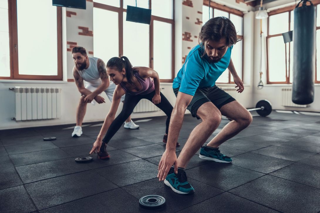 Tre persone si stanno riscaldando in una palestra con pavimento nero e grandi finestre, preparandosi per un allenamento.