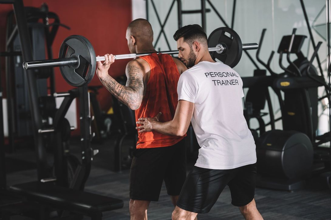 Un istruttore osserva un sollevatore di pesi mentre esegue uno squat con bilanciere in palestra.