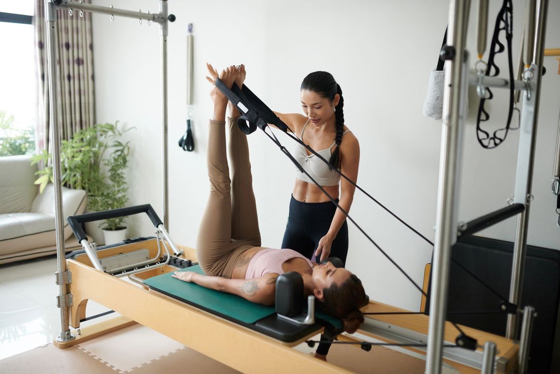 Istruttrice di Pilates assiste una cliente sul reformer con le gambe sollevate in uno studio luminoso.