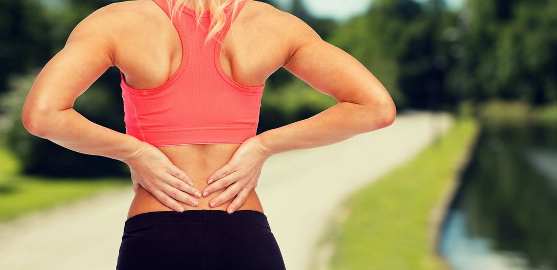 Una persona con un reggiseno sportivo color corallo si tiene la parte bassa della schiena su un sentiero esterno sfocato, segno di mal di schiena