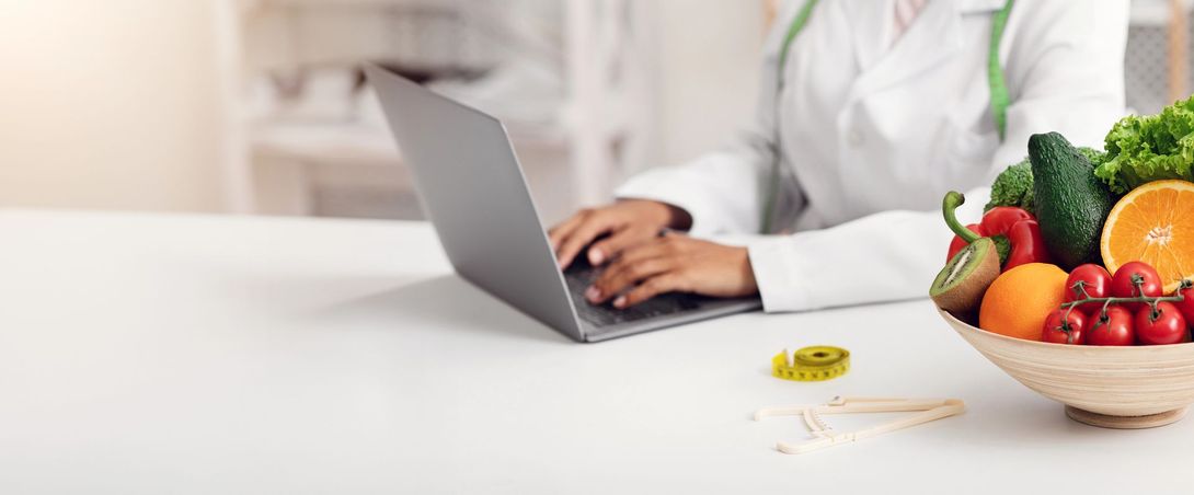 Una persona digita su un computer portatile accanto a una ciotola di frutta colorata su un tavolo bianco.