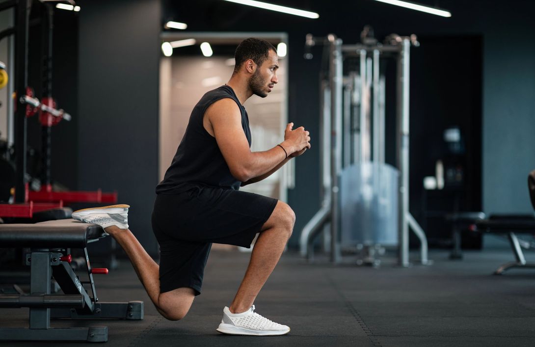 Un uomo esegue un affondo in palestra, indossando abbigliamento sportivo nero e scarpe da ginnastica bianche.