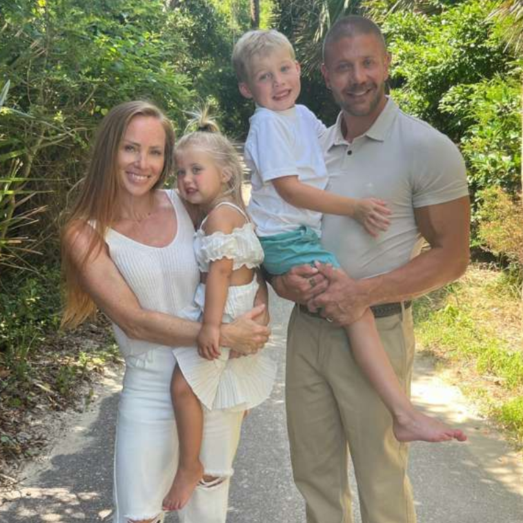 Family of four smiling outdoors on a sunny path, with two children being held by their parents.