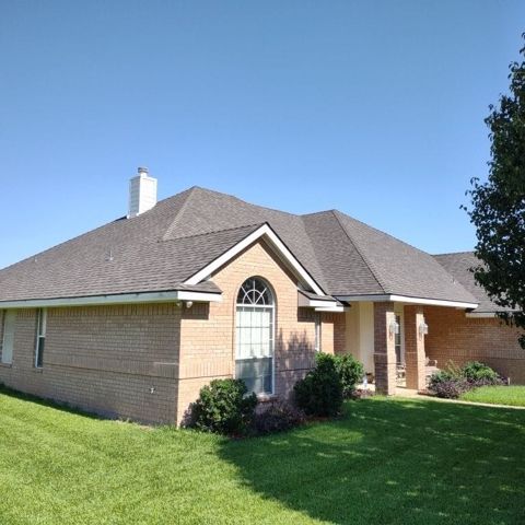 Single-story brick house with a gray roof, arched front window, and green lawn under a clear blue sky