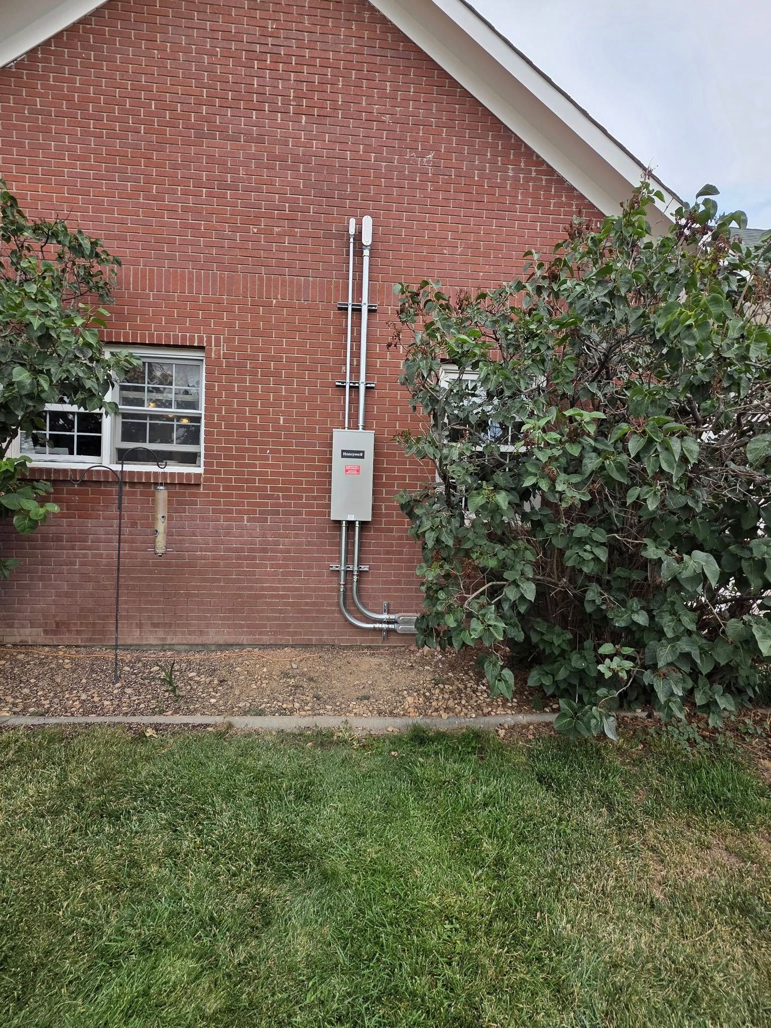 Brick house exterior with utility meter, window, and shrubs beside a grassy yard