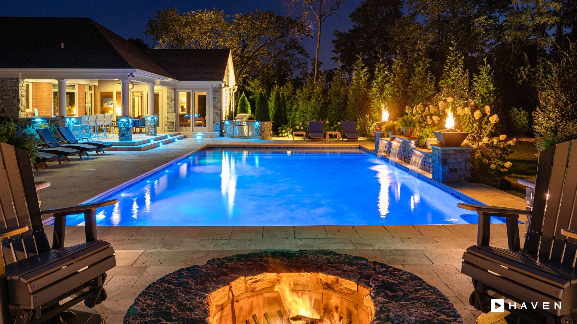 A fire pit is in front of a large swimming pool at night.