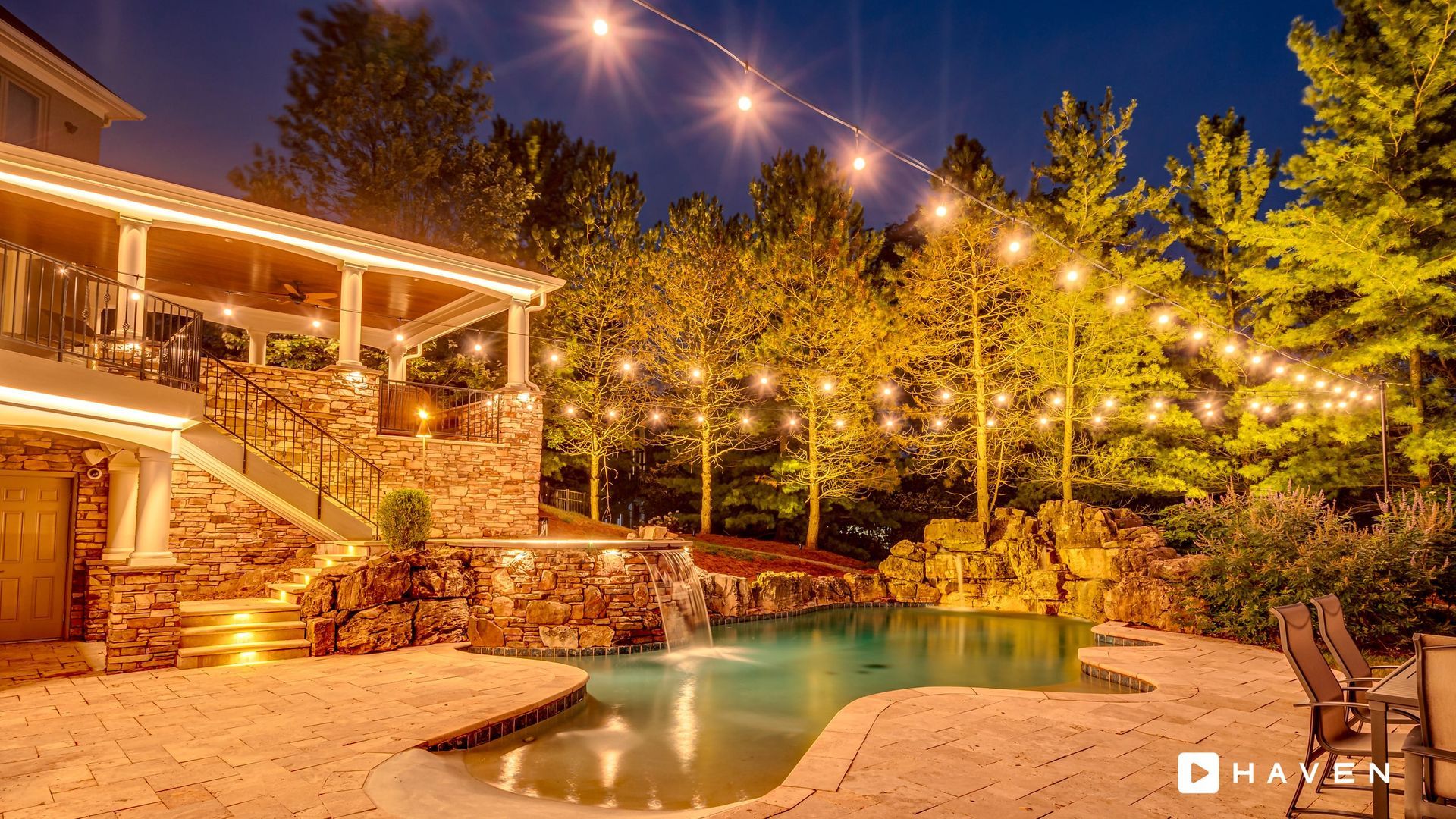 A swimming pool is lit up at night with a string of lights hanging from the trees.