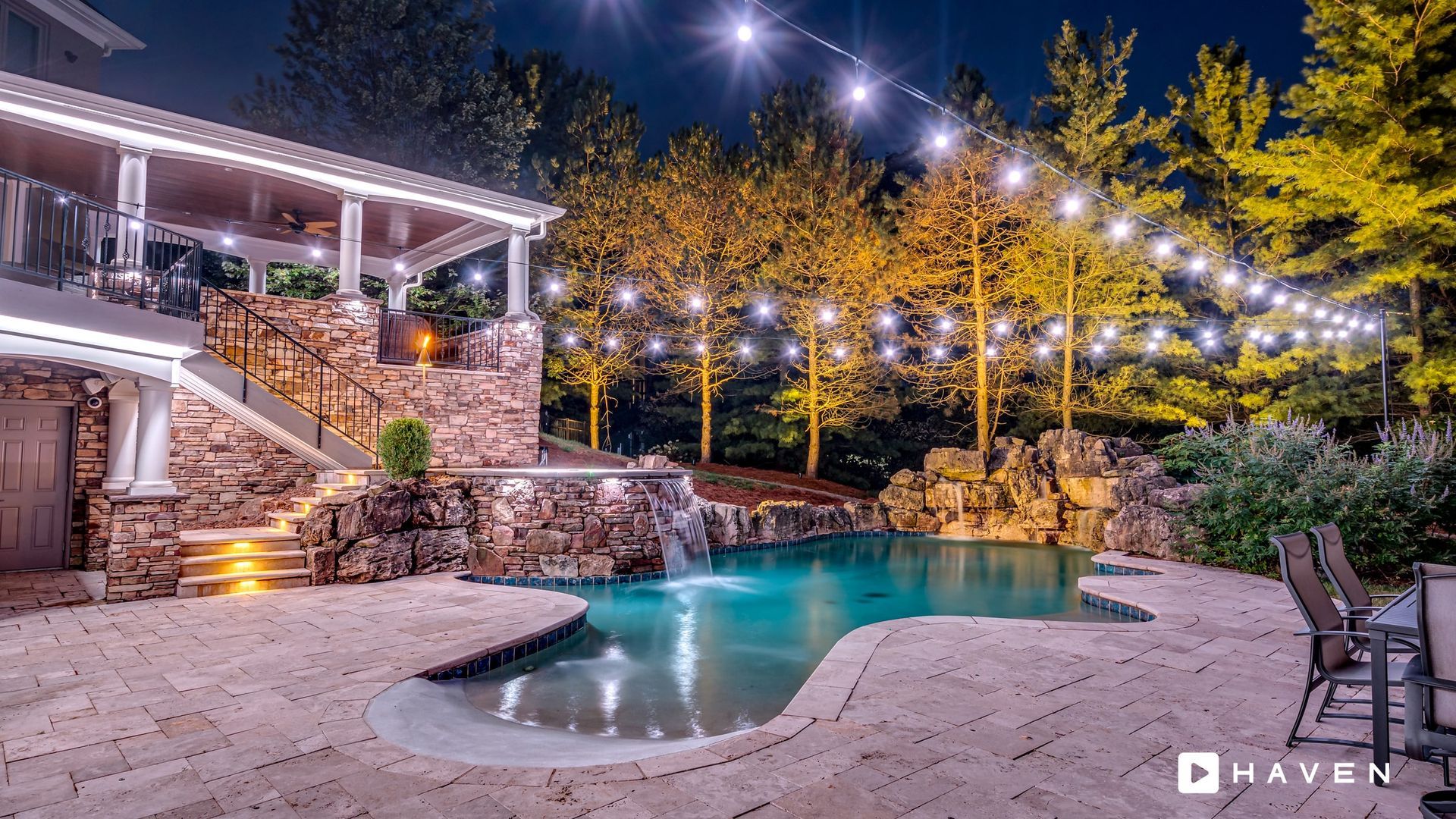 A large swimming pool is lit up at night with string lights hanging from the trees.