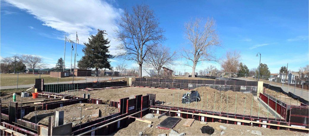 Construction site with foundation forms, trees, and buildings under a bright blue sky.