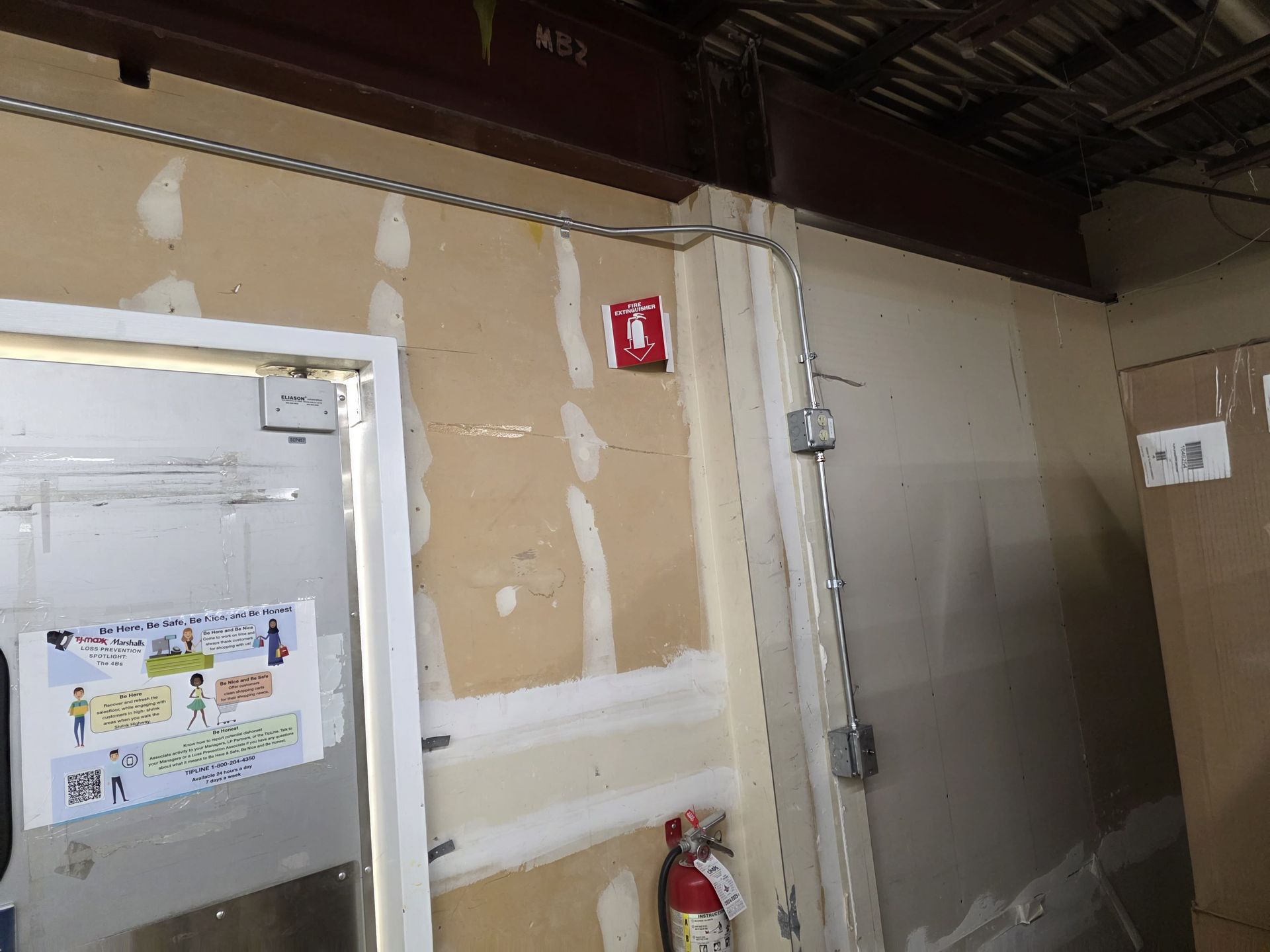 Fire extinguisher and exit signage near a doorway on a light-colored wall. Electrical conduit is visible.