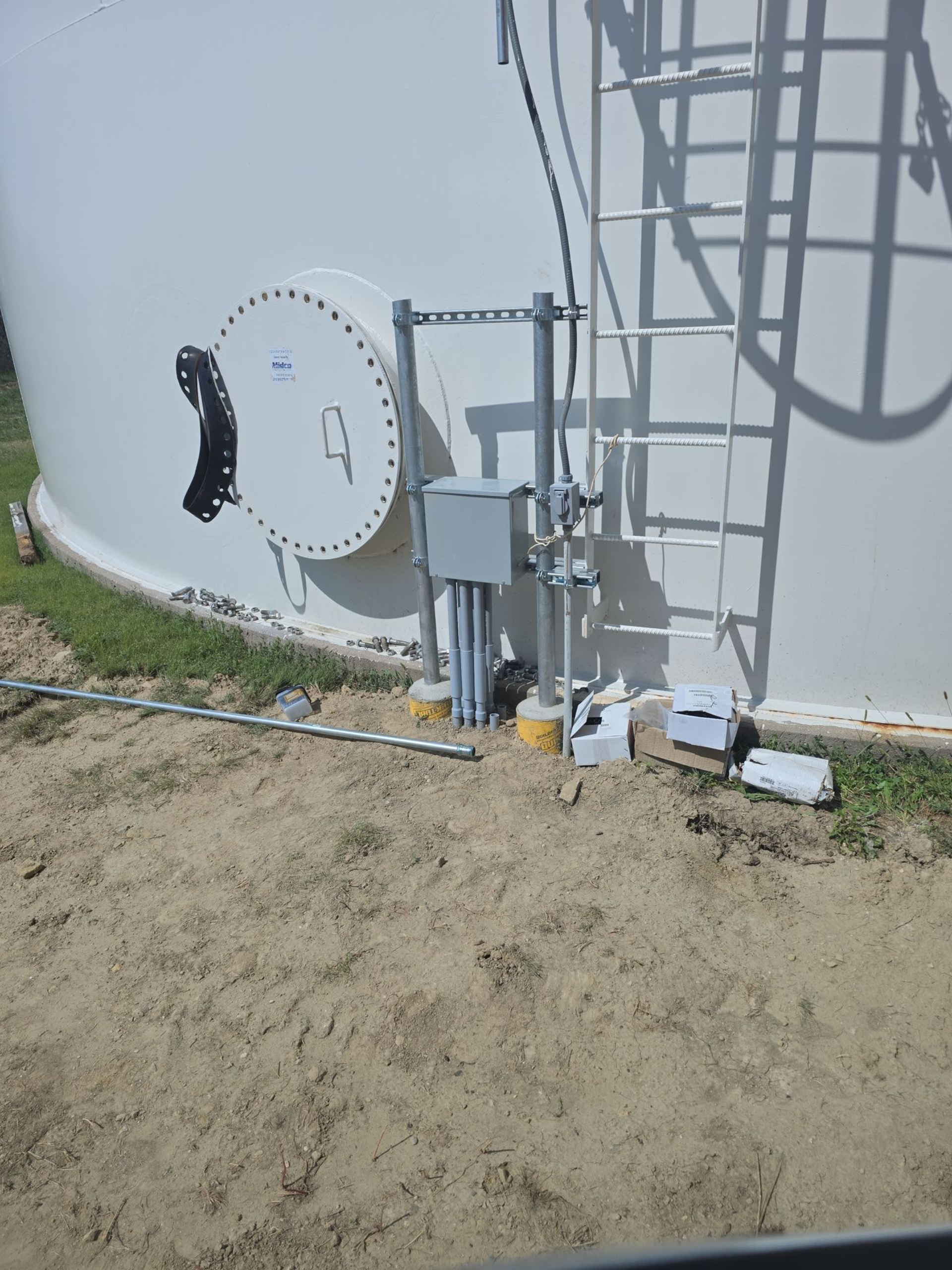 Ground-level view of a white tower with access door, ladder, and electrical box on a sandy base.