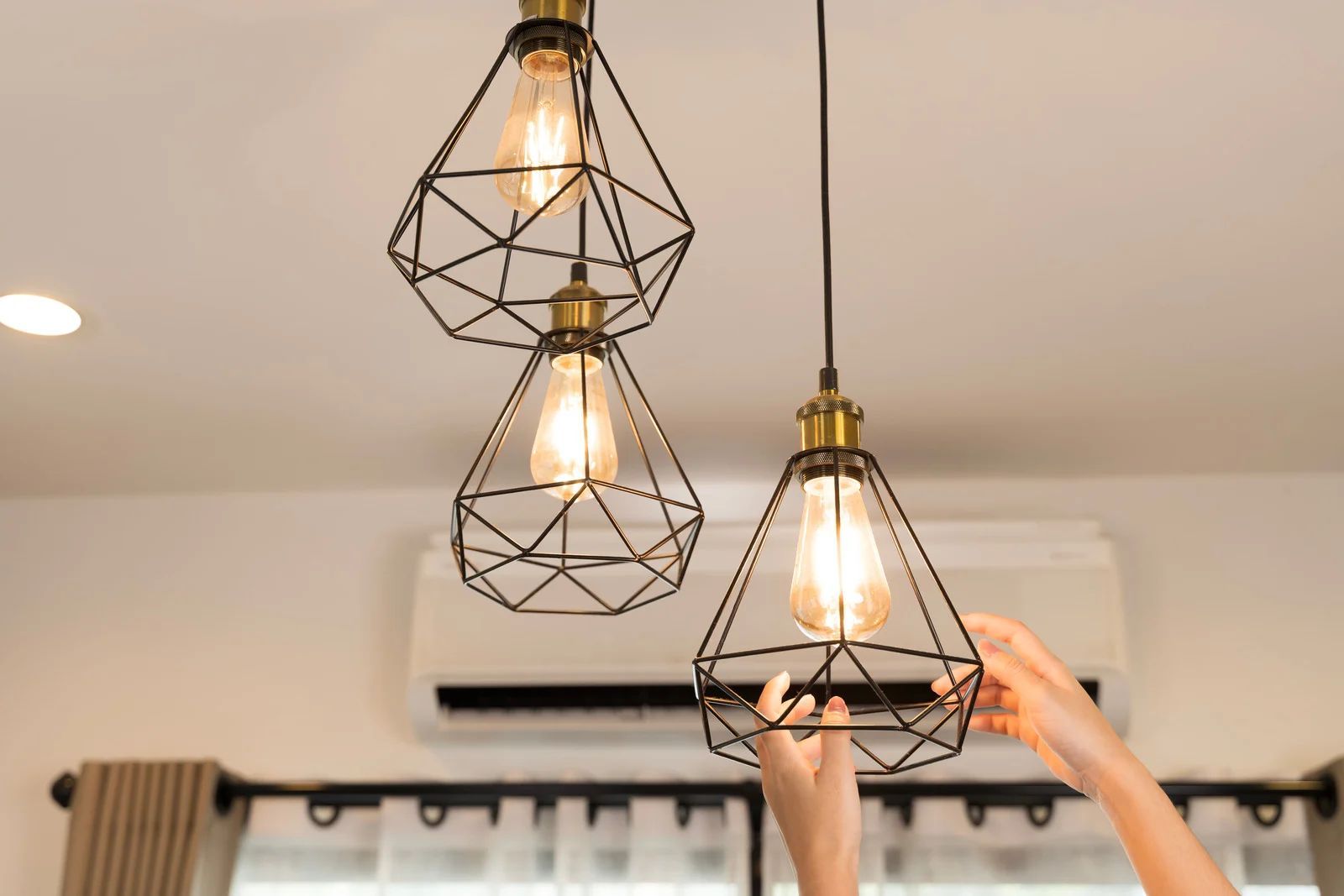 Three black geometric pendant lights hanging from a ceiling, with hands adjusting the right fixture.