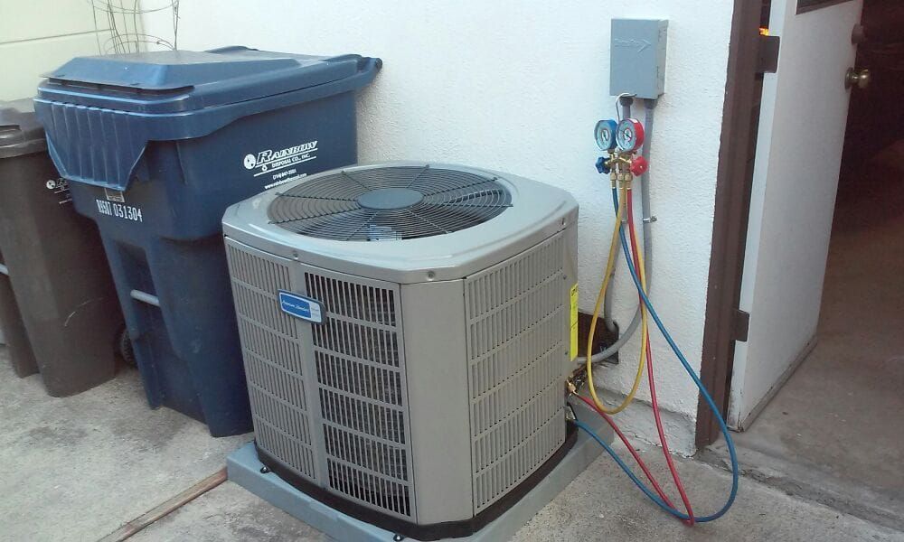 Air conditioning unit next to a blue trash bin against a white wall. Hoses and gauges are attached.