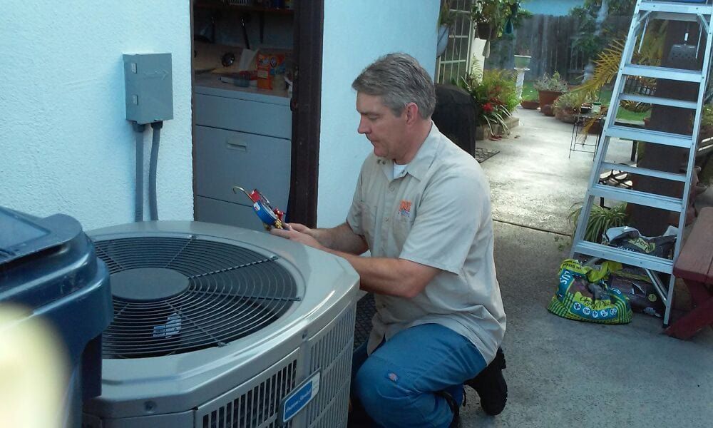Man in tan shirt kneeling, working on an AC unit outdoors. Gray unit, white wall, ladder nearby.