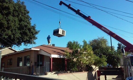 Crane lifting AC unit onto a residential roof; worker on roof guides installation under blue sky.