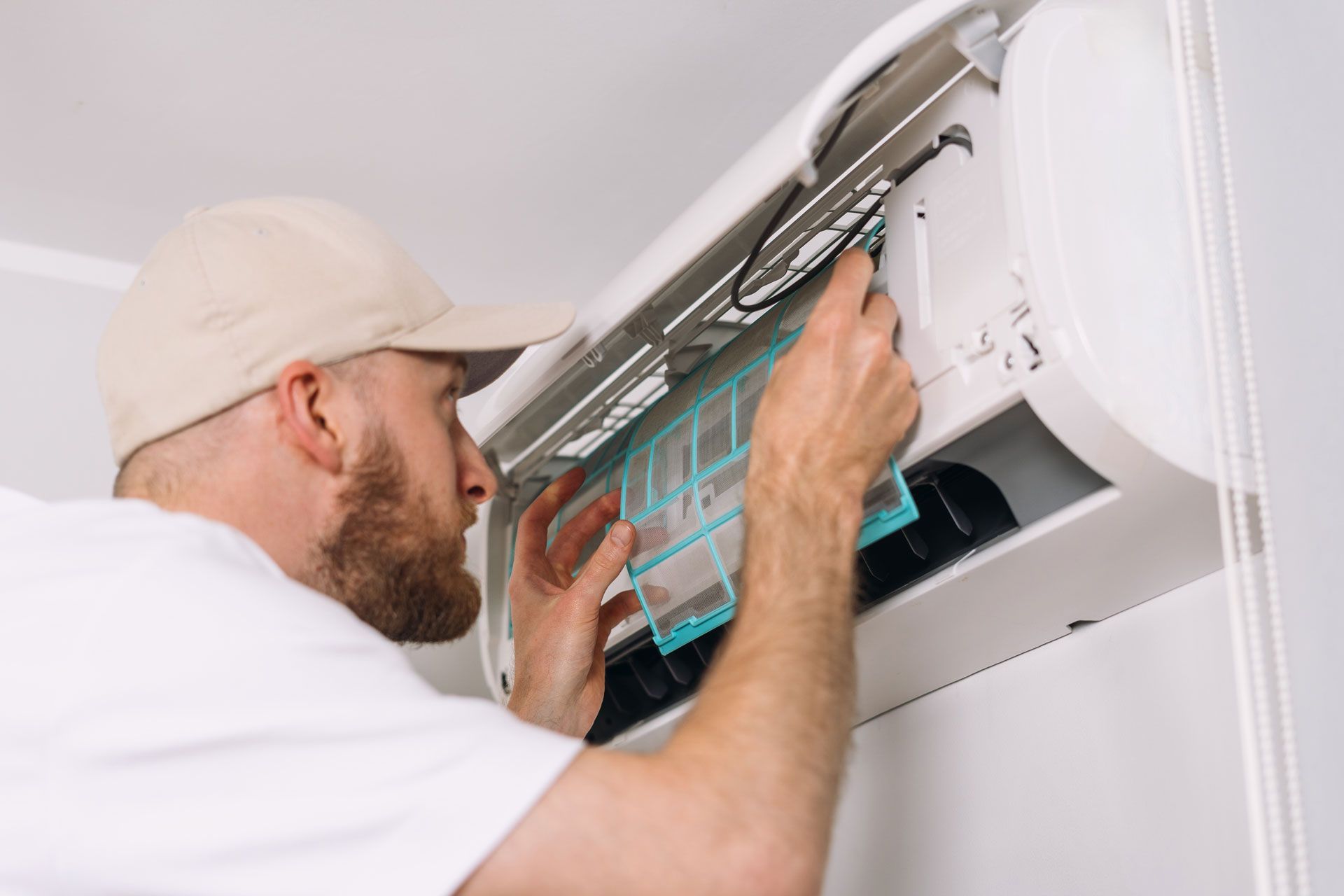 Man in cap installs air filter in a wall-mounted AC unit.