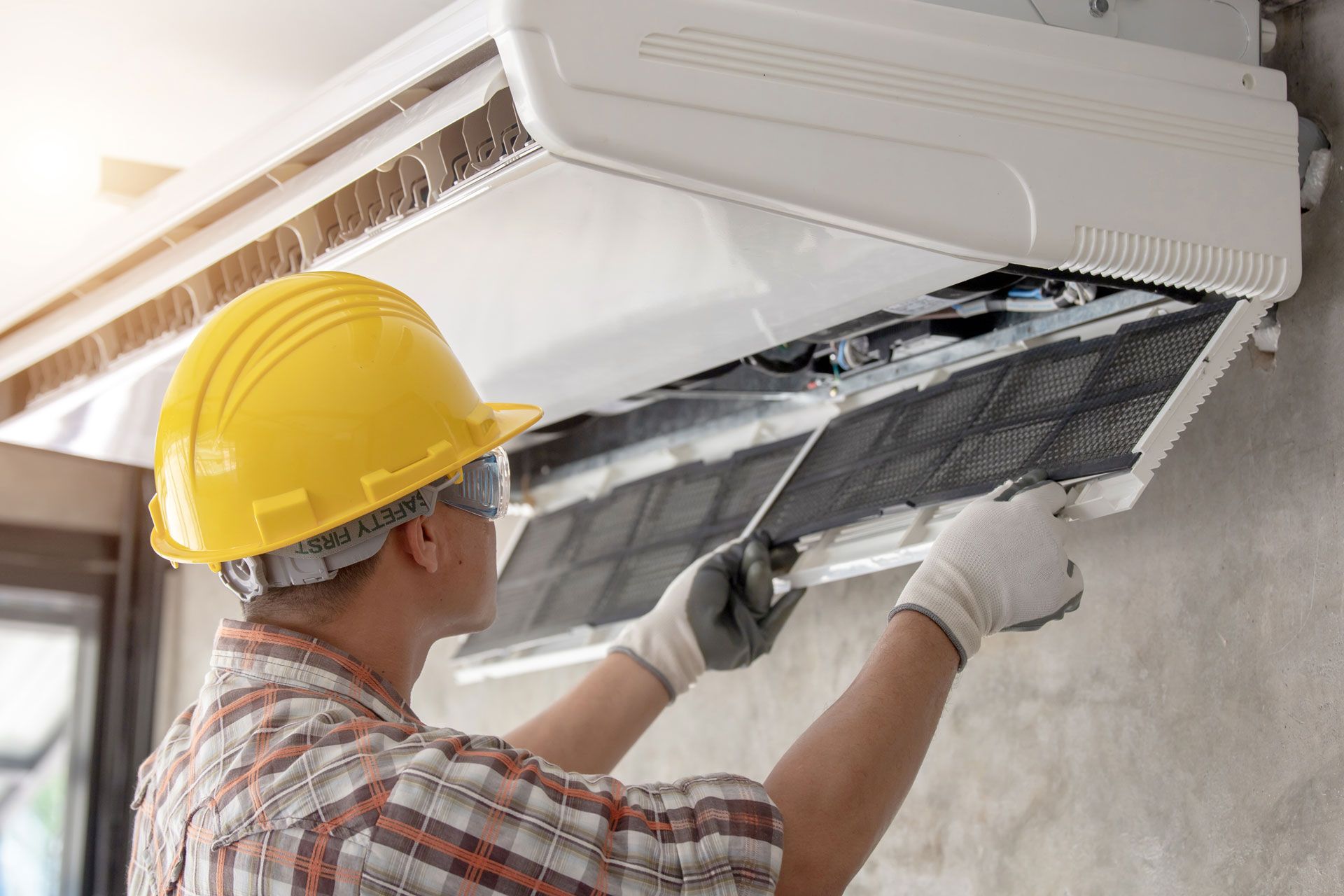 Person in a yellow hard hat and gloves cleaning an air conditioner filter.