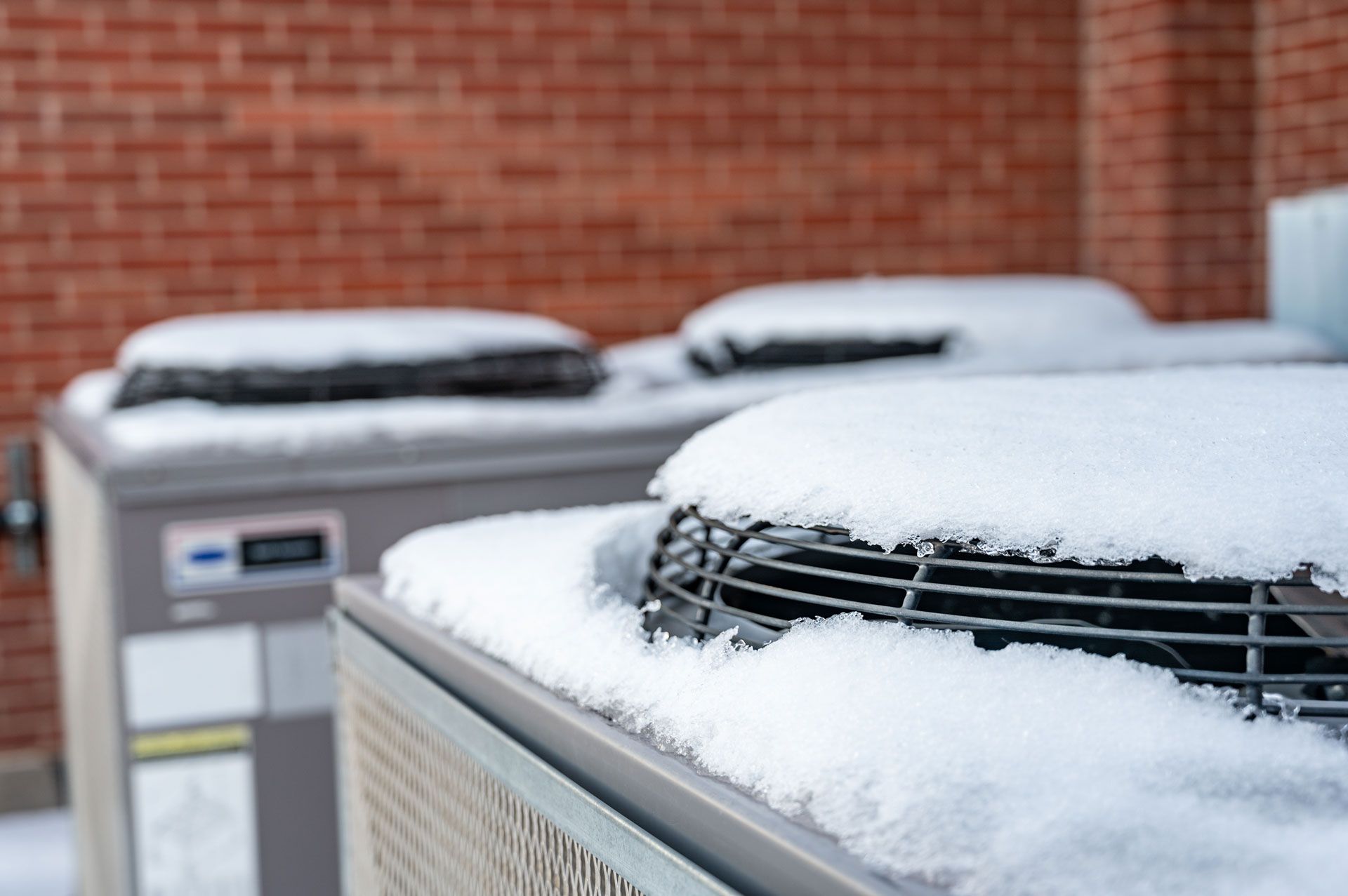 Two outdoor HVAC units covered in snow against a brick wall.