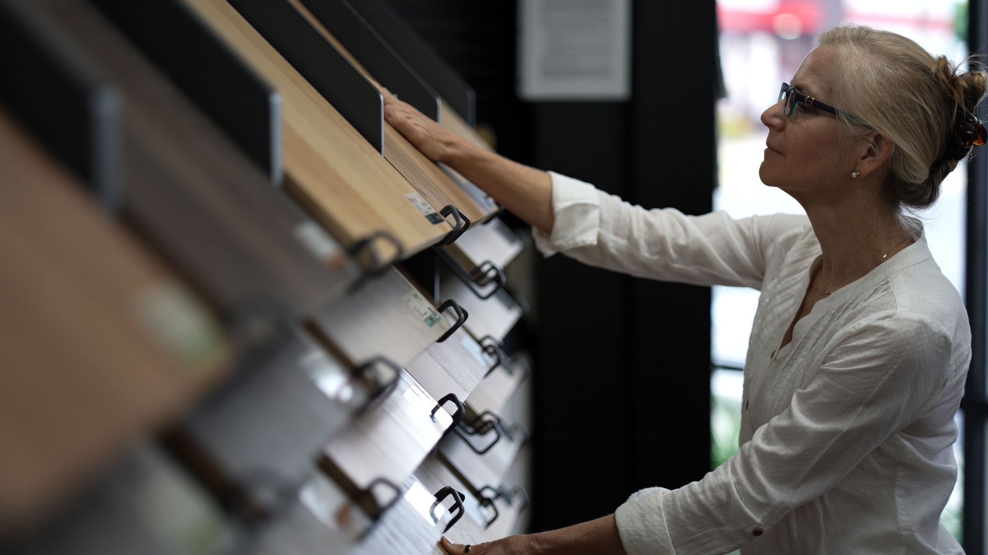 A man is showing a customer the variety of wood floors. Woman in glasses and knit hat touching samples on a wall display in a showroom
