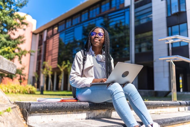 A student sitting on outdoor stairs with a laptop, smiling while looking up, with a university building in the background.