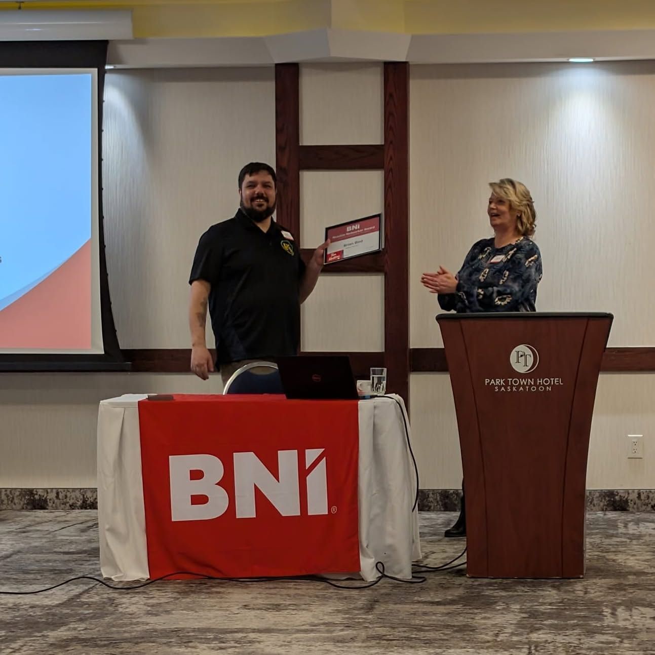 Two people on a stage beside a BNI banner and podium in a conference room