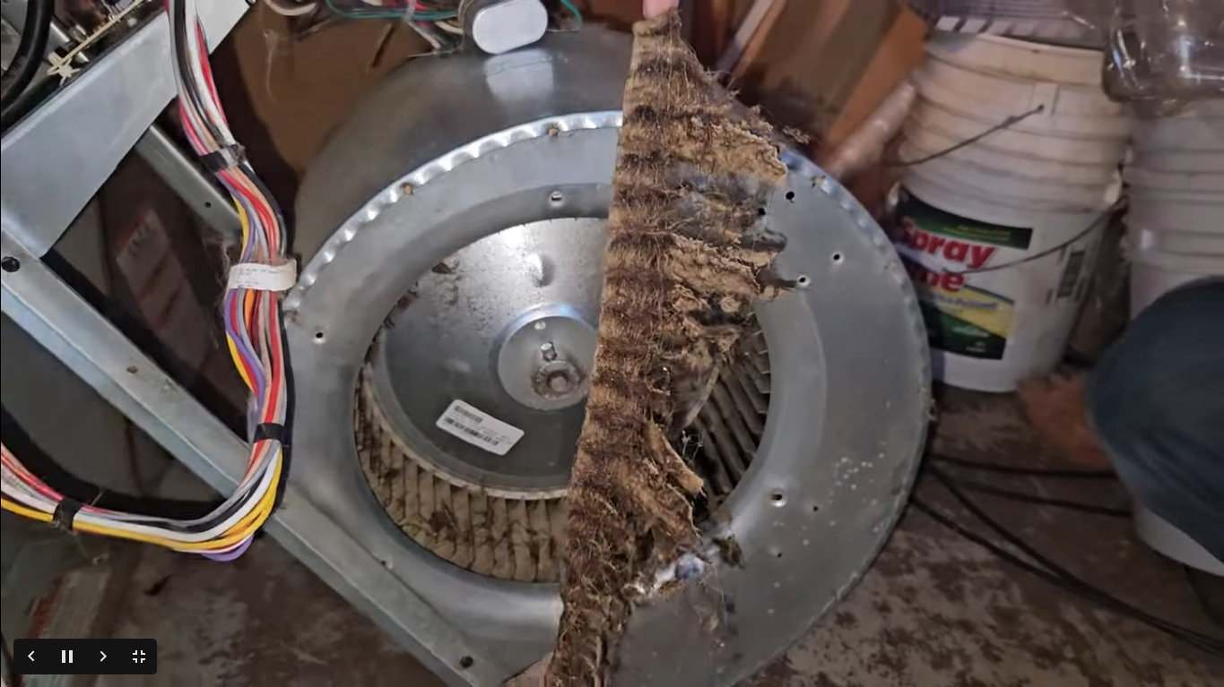 Close-up of a dusty metal fan or blower wheel with wires and debris in a cluttered indoor area