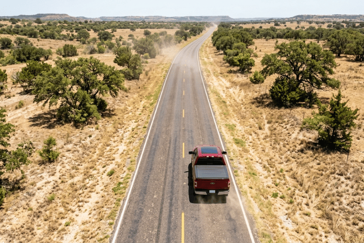 Aerial view of a pickup truck driving along a long rural highway through the Texas Hill Country with dry grasslands and scattered oak trees.
