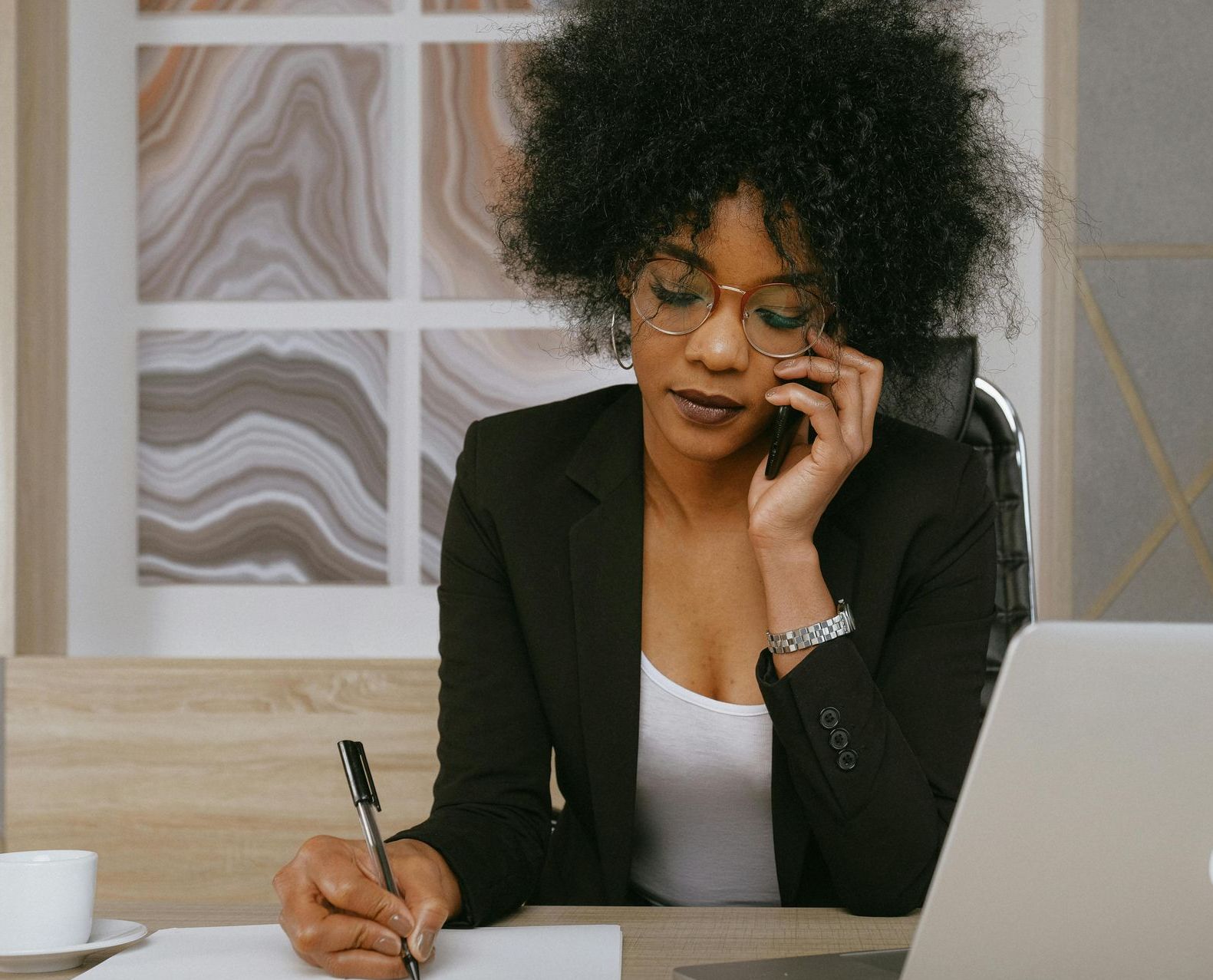 A woman is sitting at a desk talking on a cell phone and writing on a piece of paper.