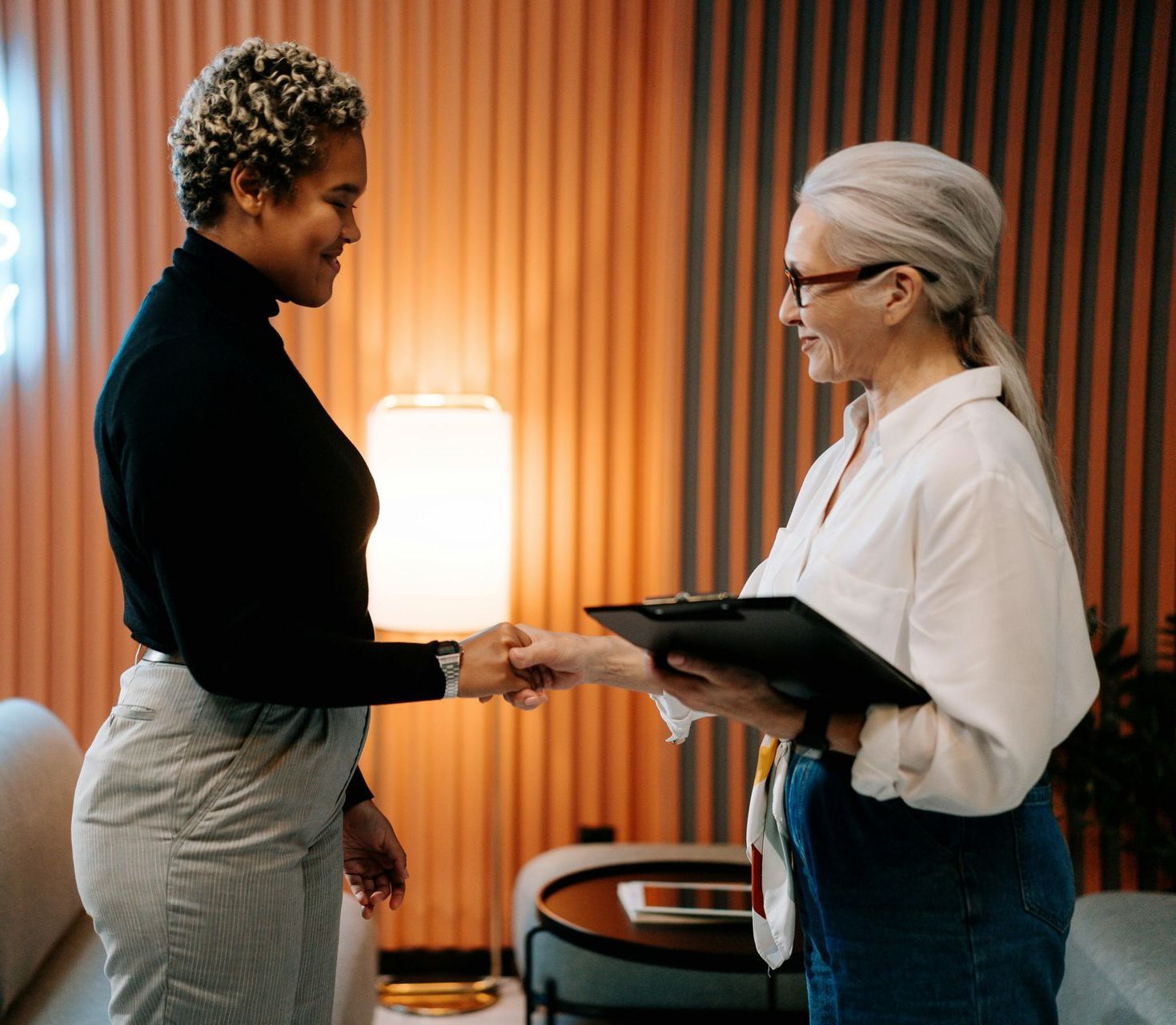 Two women are shaking hands in a living room and one is holding a clipboard.